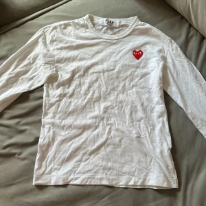Vit långärmad tröja Comme des Garçons - Säljer en vit långärmad tröja från Comme des Garçons Play med det ikoniska röda hjärtat broderat på bröstet. Tröjan är i 100% bomull och har en clean, enkel look med rund hals. Perfekt för dig som gillar stilrena plagg med en twist.