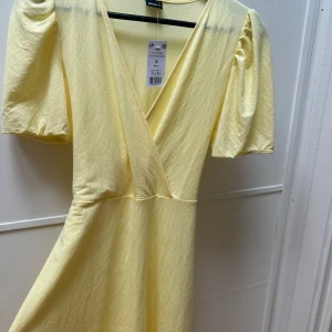 Ljusgul omlottklänning Gina  - Supersöt ljusgul omlottklänning från Gina med puffiga korta ärmar och v-ringning. Klänningen har knytband i midjan och är tillverkad i mjuk bomull, perfekt för sommarens alla tillfällen. Prislapp kvar.