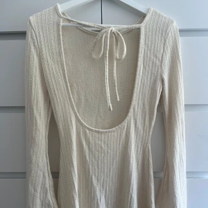 Ribbmönstrad vit långärmad klänning - Supersnygg långärmad klänning i ribbad struktur med diskret mönster. Djup urringning i ryggen med knytband upptill. Färgen är krämvit och materialet känns mjukt och stretchigt, perfekt som en sommarklänning.