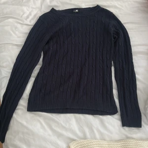 Mörkblå kabelstickad tröja JC - Mörkblå kabelstickad tröja från JC med rund halsringning och långa ärmar. Tröjan har ribbade muddar vid ärmslut och nederkant. Den är i storlek L men skulle kunna passa en M