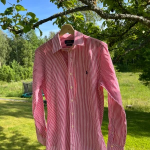 Rödvit randig skjorta Polo Ralph Lauren - Snygg röd och vit randig skjorta från Polo Ralph Lauren i slim fit. Skjortan har klassisk krage, långa ärmar och knäppning framtill. Broderad blå logga på bröstet. Tillverkad i stretchig bomull för extra komfort. Perfekt nu till sommaren!