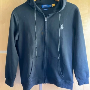 Svart hoodie från Polo Ralph Lauren - Svart hoodie från Polo Ralph Lauren i 100% bomull med dragkedja framtill, huva med snörning och klassisk broderad logga på bröstet. Två fickor framtill och ribbade muddar. Snygg och enkel att matcha till det mesta. Storlek M men funkar även som S.                  Hör av dig vid frågor eller för fler bilder! 
