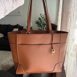 Michael Kors väska - Snygg brun axelväska från Michael Kors i stilren design med guldiga detaljer och logotyp. Väskan är tillverkad i skinn och har flera fack, både invändigt och utvändigt, samt dragkedja upptill. Perfekt för dig som vill ha plats för allt du behöver.