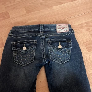 True Religion jeans - Snygga blå jeans från True Religion med klassisk bakficka och kontrastsömmar. Jeansen har en ikonisk logga på bakfickan och patch bak i midjan. De är i modellen Joey och är lågmidjade