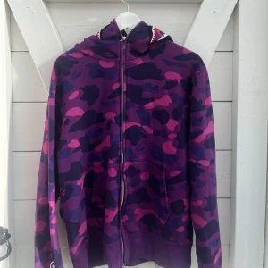 BAPE Color Camo Shark Full Zip Hoodie - Säljer en ikonisk BAPE hoodie i lila och rosa camo med full zip och haj-tryck på huvan. Tröjan har broderade bokstäver, tiger-motiv och klassiska kängurufickor. Perfekt statement-plagg för dig som gillar streetwear och unika detaljer. Kvitto och stock X tag fixas till intresserad köpare