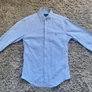 Blårandig skjorta från Ralph Lauren - Snygg blårandig skjorta från Ralph Lauren i slim fit-modell. Skjortan har klassisk krage, långa ärmar och knäppning framtill. Tillverkad i mjuk bomull och prydd med den ikoniska broderade loggan på bröstet. Perfekt för dig som gillar stilrena och tidlösa plagg.