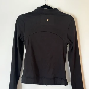 Svart träningsjacka - Svart, figursydd träningsjacka med hel dragkedja framtill och två fickor med dragkedja. Jackan har hög krage och är tillverkad i ett stretchigt, mjukt material som passar perfekt till träning eller aktiva dagar. Den köptes secondhand därav vet jag inte om den är äkta eller inte.