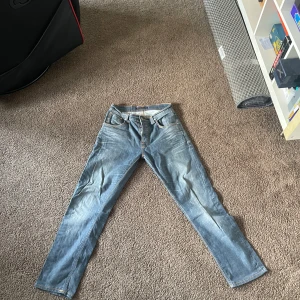Ljusblå jeans från Nudie Jeans - Säljer ett par ljusblå jeans från Nudie Jeans med klassisk femficksdesign och orange kontrastsömmar. Jeansen har rak passform och är tillverkade i mjukt denimtyg. Snygga slitningar framtill och broderade bakfickor ger en cool look.