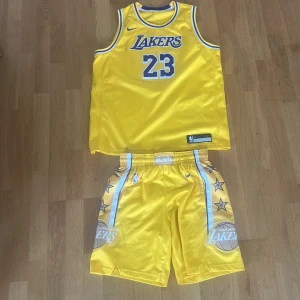 Gul Lakers basketlinne LeBron James #23 - Säljer ett gult basketlinne från Nike med Lakers-tryck och nummer 23, LeBron James. Linnet har blå och vita detaljer, är ärmlöst och tillverkat i polyester. Perfekt för basket eller sportiga outfits. Matchande shorts med stjärnor och Lakers-logga ingår. Givetvis äkta och införskaffade på basketshop för x antal år sedan. Storleken är XL i barnstorlek men passar S-M beroende på hur man gillar sin passform. 
