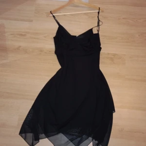 Svart asymmetrisk klänning Pipeline - Svart kortklänning från Pipeline med tunna axelband och v-ringad urringning. Klänningen har ett lager av tunn, transparent polyester som ger en snygg asymmetrisk och flowy nederdel. Perfekt för dig som gillar en stilren men edgy look.