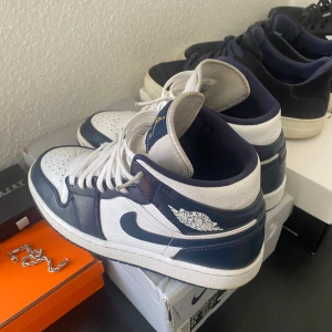  Jordan 1 Mid Navy/Vit - Säljer ett par Nike Air Jordan 1 Mid i marinblått och vitt skinn. Klassisk siluett med högt skaft, perforerad tå och ikoniska Air Jordan-loggan på sidan. Skorna har vita snören och detaljer i guld. Storlek 40,5 men passar som 41, kommer även med ett par blåa snören med! 