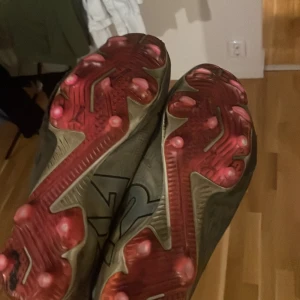 Gråa fotbollsskor från Puma - Säljer ett par gråa fotbollsskor från Puma med snörning och röda dobbar undertill. Skorna har en sportig design med Pumas logga på ovansidan och är tillverkade i syntetmaterial för bästa grepp på planen. De är elite 