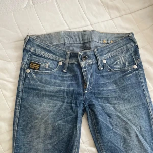 Blå jeans från G-Star RAW - Säljer ett par blå jeans från G-Star RAW med snygg slitning och klassiska fem fickor. Jeansen har låg midja, raka ben och coola detaljer som broderad logga bak och slitna partier. Midjemått 39 rakt över, 77 i innerberndlängd 