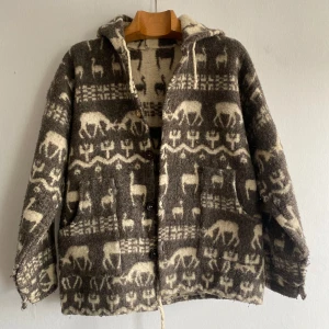 Mönstrad ulljacka med huva - Unik jacka i tjock ull med brunt och beige mönster av renar o h lamor och geometriska former. Jackan har huva, stora fickor och knäpps med träknappar framtill, knappar finns på insidan oxå. Snöre längst ner så du kan spänna åt/justera längden. Perfekt för dig som gillar boho-vibe och vill sticka ut med ett cozy plagg. Sista bilden visar en liten mini missfärgning. Jackan uppskattas till en Large.