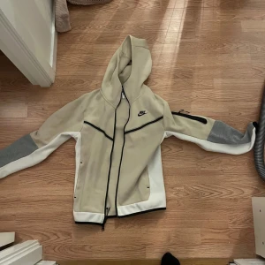 Beige och grå hoodie från Nike. (Nike tech)  - Snygg hoodie från Nike i beige och grått med svarta detaljer. Hoodie har en dragkedja framtill och en praktisk huva. Perfekt för en sportig look. Nike tech 