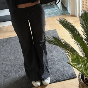 Svarta randiga kostymbyxor bootcut - Snygga svarta kostymbyxor med tunna vita ränder och bootcut-silhuett. Byxorna har låg midja och är i ett klassiskt kostymtyg som känns dressat men ändå chill. Perfekta att styla med sneakers eller boots för en cool look.