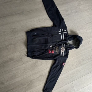 Svart hoodiejacka Geographical Norway S - Svart hoodiejacka från Geographical Norway med broderade detaljer, norska flaggor och patchar. Jackan har huva, dragkedja, fickor och en extra ficka på ärmen. Snygga vita och gråa detaljer samt brodyr framtill ger en sportig vibe.priset är ej hugget i sten 