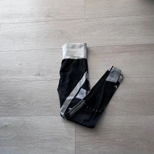 Svarta och gråa träningstights med mönster - Säljer ett par svarta träningstights med grått och vitt grafiskt mönster och bred, ribbad midja. Snygga detaljer längs benen och stretchigt material som sitter tight. Perfekta för gymmet eller löpning.