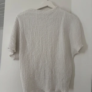 Vit topp  - Säljer en vit topp med ribbad struktur och korta ärmar. Tröjan har en avslappnad passform och är tillverkad i ett mjukt material som känns skönt mot huden. Perfekt för en enkel och stilren look.