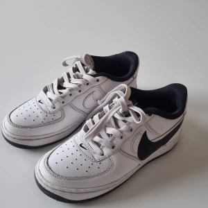 Vita Nike Air Force 1 sneakers - Klassiska vita Nike Air Force 1 sneakers med svart swoosh och svarta detaljer. Skorna har snörning, perforerade detaljer på tån och en platt sula. Perfekta för dig som gillar en tidlös och sportig stil.