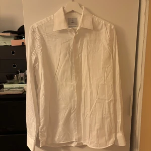 Vit skjorta från Bläck - Klassisk vit skjorta från Bläck i modellen Tailored Fit. Skjortan har lång ärm, knappar framtill och en stilren krage. Perfekt för dig som vill ha en enkel och snygg skjorta till garderoben.