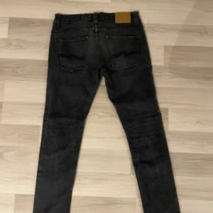 Svarta jeans från Nudie Jeans - Snygga svarta jeans från Nudie Jeans med klassisk femficksdesign. De har en knappgylf och subtila orangea detaljer vid fickorna. Perfekta för en stilren look.