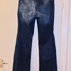 Mörkblå bootcut jeans med knappar - Snygga mörkblå jeans med bootcut passform och slitningar framtill. Jeansen har dubbla knappar i midjan och klassiska fickor både fram och bak. Perfekta för en avslappnad och trendig look.