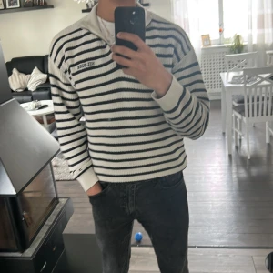 Vit och marinblå randig stickad tröja från Hilfiger - Säljer en stickad tröja från Hilfiger i vitt med marinblå ränder. Tröjan har halv dragkedja vid kragen och broderad logga på bröstet. Klassisk passform och långa ärmar, perfekt för en stilren look.