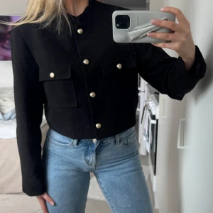 Svart croppad kavaj från NA-KD - Säljer en svart croppad kavaj från NA-KD med guldknappar och två fickor framtill. Jackan har lång ärm och en stilren, rak passform. Nypris 699. 