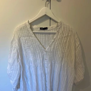 Vit blus med knappar från Cubus - Säljer en luftig vit blus från Cubus i storlek 42. Blusen har v-ringning, knappar framtill och ett diskret randigt mönster i tyget. Perfekt för en avslappnad och stilren look. Den sitter som en M/L