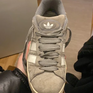 Grå Adidas Campus sneakers - Säljer ett par klassiska Adidas Campus sneakers i grå mocka med vita detaljer och snörning. Skorna har den ikoniska trefoil-loggan på plösen och hälen samt vita ränder på sidan. Perfekta för en stilren vardagslook.