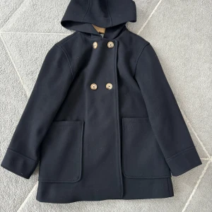 Marinblå kappa med huva och knappar - Säljer en marinblå kappa med huva och stora fickor framtill. Kappan har dubbelknäppning med fyra ljusa knappar och långa ärmar. Perfekt för våren och enkel att matcha med olika outfits. Stor i storleken. Pris kan diskuteras!