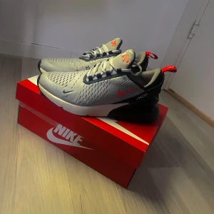 Nike Air Max 270 sneakers i grått och svart - Nike Air Max 270 sneakers i grått och svart med röda detaljer och synlig Air Max-sula. Ovandelen är i mesh med svarta och grå paneler, röda loggor och dragflik bak. Snygg och sportig modell med snörning och platt sula. Den har lite defekt som inte märks
