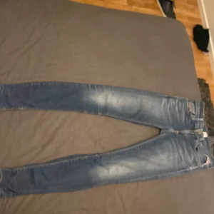 Blå Replay jeansbyxor med slitningar - Säljer ett par blå jeansbyxor från Replay med snygga slitningar och klassisk femficksdesign. Byxorna har normal passform och är tillverkade i denim med Replay-logga på insidan av linningen. Perfekta för dig som gillar en tidlös jeanslook.