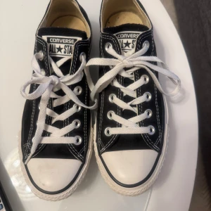 Svarta Converse All Star sneakers - Klassiska svarta Converse All Star sneakers med vit sula och vita snören. Skorna har rund tå, platt sula och tydliga vita sömmar. Perfekta för en avslappnad och tidlös stil.