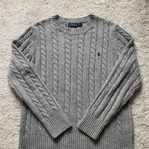 Kabelstickad tröja från Ralph Lauren - Säljer en klassisk grå kabelstickad tröja från Polo Ralph Lauren med rund halsringning och broderad logga på bröstet. Tröjan har långa ärmar och breda ribbade muddar vid ärmslut och nederkant. Perfekt för lager-på-lager under kyliga dagar. (Bara använd några enstaka gånger).