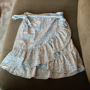 Blommig omlottkjol i blått från ONLY - Supersöt blå omlottkjol från ONLY med vitt blommönster och volangdetaljer. Kjolen har knytband i midjan och är perfekt för en somrig look. Lätt och luftig modell som ger en feminin känsla.