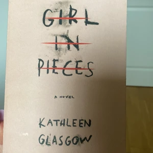 Girl in Pieces - Säljer boken 'Girl in Pieces' av Kathleen Glasgow. Omslaget är ljusrosa med svart handskriven text och röda överstrykningar. Romanen är på engelska och handlar om Charlotte Davis och hennes kamp med svåra känslor och livshändelser.