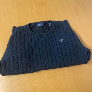 Mörkblå kabelstickad tröja från GANT - Säljer en klassisk mörkblå kabelstickad tröja från GANT med rund halsringning och diskret logga på bröstet. Tröjan har långa ärmar och ribbade muddar vid ärmslut och nederkant. Perfekt för en stilren och tidlös look.
