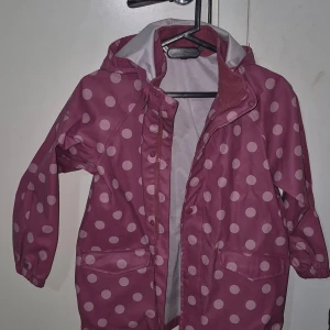 Rosa prickig regnjacka från H&M - Rosa regnjacka från H&M med vita prickar och huva. Jackan har dragkedja framtill, två fickor och är helfodrad med ljusgrått tyg. Perfekt för blöta dagar när du vill sticka ut lite extra.