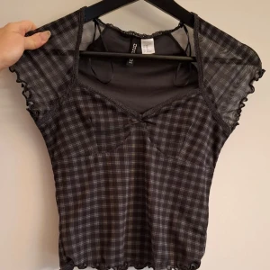 Svart och grå rutig topp från H&M Divided - Säljer en svart och grå rutig topp från H&M Divided i storlek XS. Toppen har korta ärmar, spetsdetaljer vid halsringningen och en liten rosett framtill. Tunt och transparent tyg.