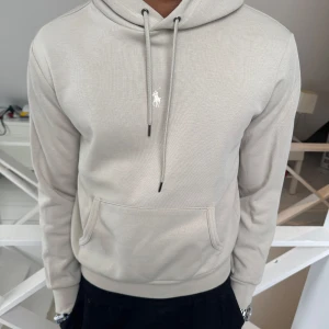 Beige hoodie från Ralph Lauren - Säljer en stilren beige hoodie från Ralph Lauren med klassisk broderad logga på bröstet. Tröjan har huva med dragsko och en stor magficka framtill. Perfekt för en avslappnad och snygg look.
