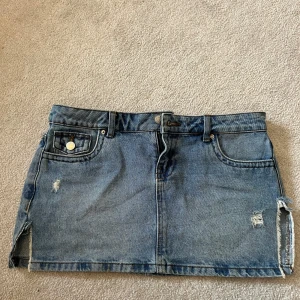 Blå jeanskjol från Noisy May - Säljer en lågmidjad blå jeanskjol från Noisy May med fem fickor, slitna detaljer och råa kanter. Kjolen har en kort passform och dragkedja fram. Perfekt för dig som gillar en avslappnad och trendig stil. Midjemåttet är 41 och rakt ner på mitten: 29.5 cm.