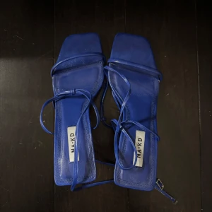 Blå klackade skor från NA-KD - Säljer ett par blå klackar från NA-KD med smala remmar och fyrkantig tå. Skorna har klack och långa snören som kan lindas runt vristen. Dom är helt oanvända som bara legat i skåpet och dammar, dom förtjänar nytt hem🥰 passar dig som är 37 och 38