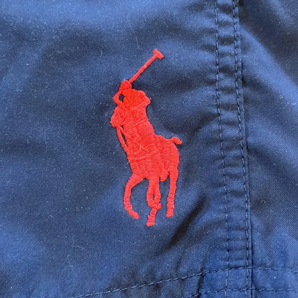 Snygga marinblå badshorts från Polo Ralph Lauren med röd broderad logga på benet och röd snörning i midjan. Klassisk modell med normal passform, perfekt för sommaren. Tillverkade i polyester för snabb torkning. Storlek: M (10-12). Hör av dig vid frågor!. Muu.
