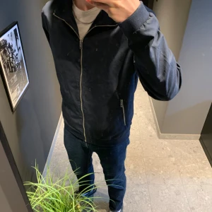 Mörkblå jacka från Jack & Jones - Snygg mörkblå bomberjacka från Jack & Jones med dragkedja framtill och två fickor med dragkedjor. Jackan har en klassisk krage och långa ärmar. Perfekt för en stilren look. 