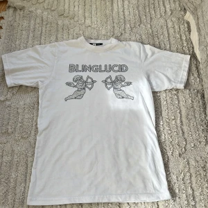 Vit t-shirt med tryck från Blinglucid - Vit t-shirt från Blinglucid med tryck av två änglar och texten 'BLINGLUCID' framtill. Klassisk passform och rund halsringning. Perfekt för en avslappnad och trendig stil.
