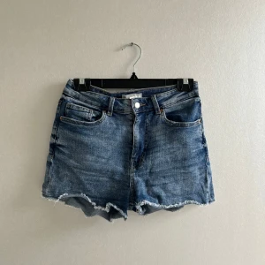 Blå jeansshorts med slitningar längs kanterna  - Säljer ett par blå jeansshorts med fransig, rå kant och femficksmodell. Shortsen har knapp och dragkedja framtill samt bälteshällor. Perfekta för sommarens outfits.