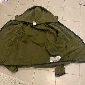 Olivgrön hoodiejacka från Nike - Säljer en olivgrön hoodiejacka från Nike med dragkedjeficka på ena ärmen och svarta kantdetaljer. Jackan har huva och är långärmad, perfekt för träning eller chill. Snygg och enkel design med sportig känsla.