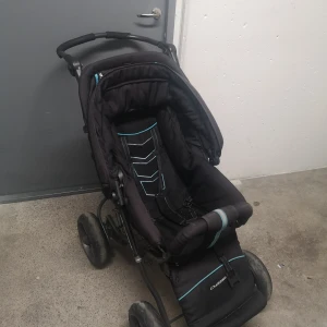 Svart barnvagn Crescent - Säljer en svart barnvagn från Crescent med turkosa och vita detaljer. Vagnen har fyra stora hjul, justerbart handtag och en rymlig varukorg undertill. Sitsen är vadderad och har sele för säkerhet. Perfekt för promenader i stan eller parken.
